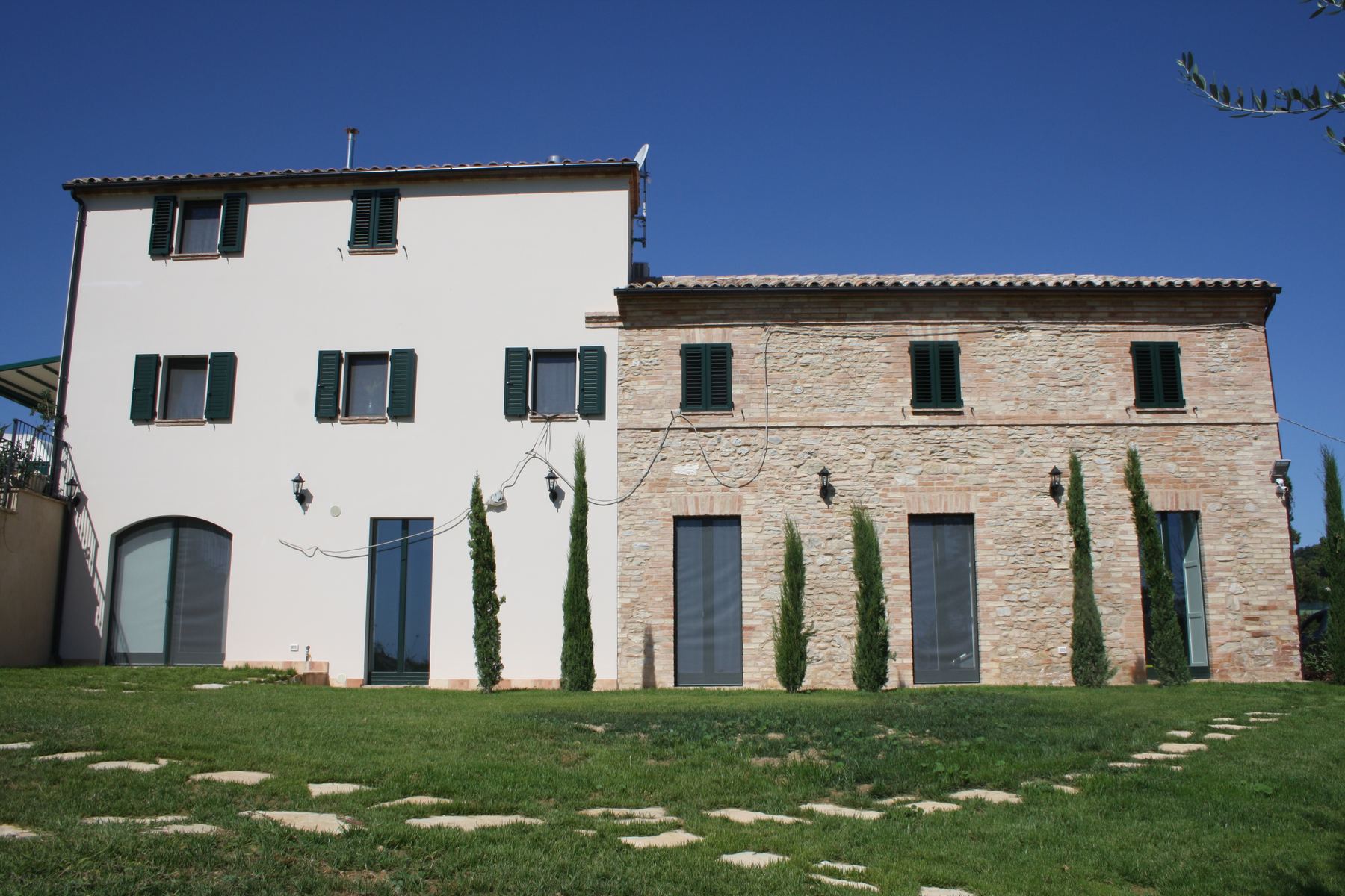 Prospetto lungo di un casale, metà intonacata di bianco e metà in pietra, con cipressi allineati sul prato
