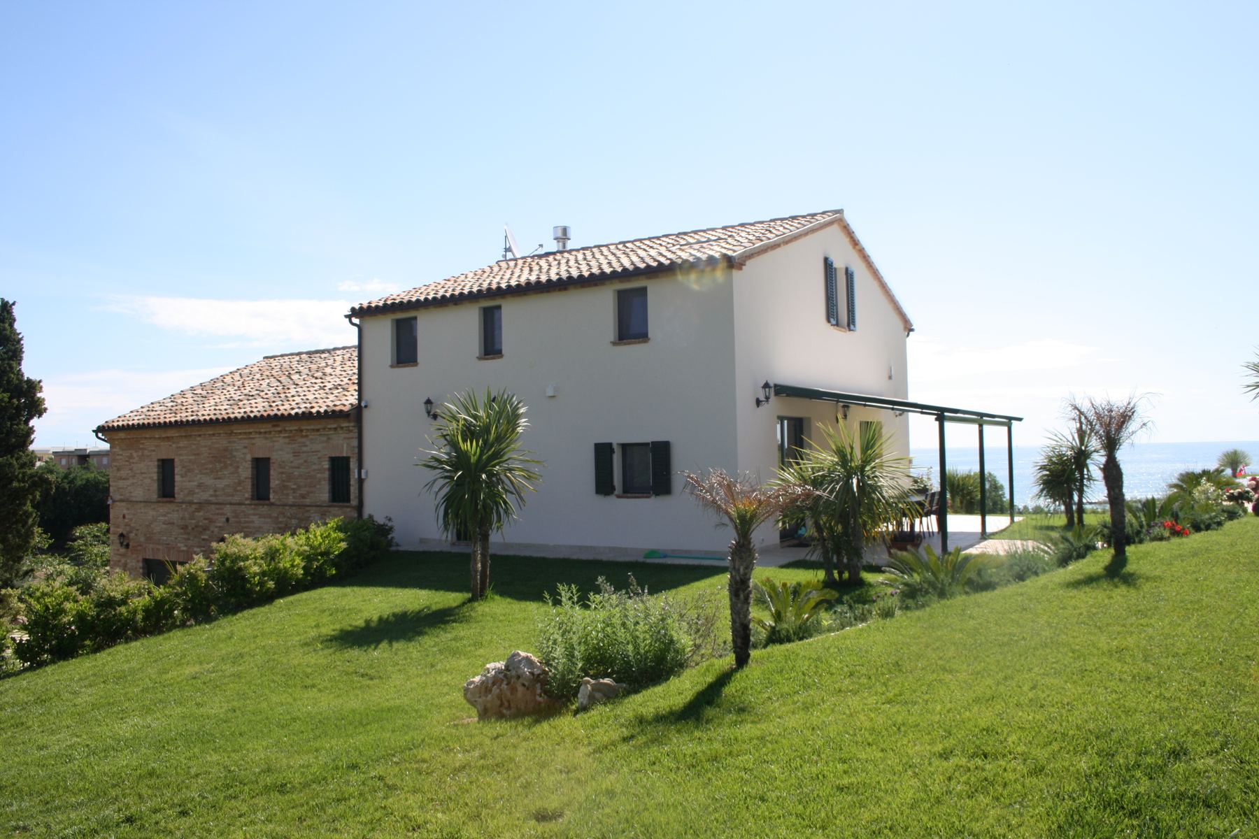 Casale bianco con ala in pietra su un prato in pendenza, palme e mare all'orizzonte