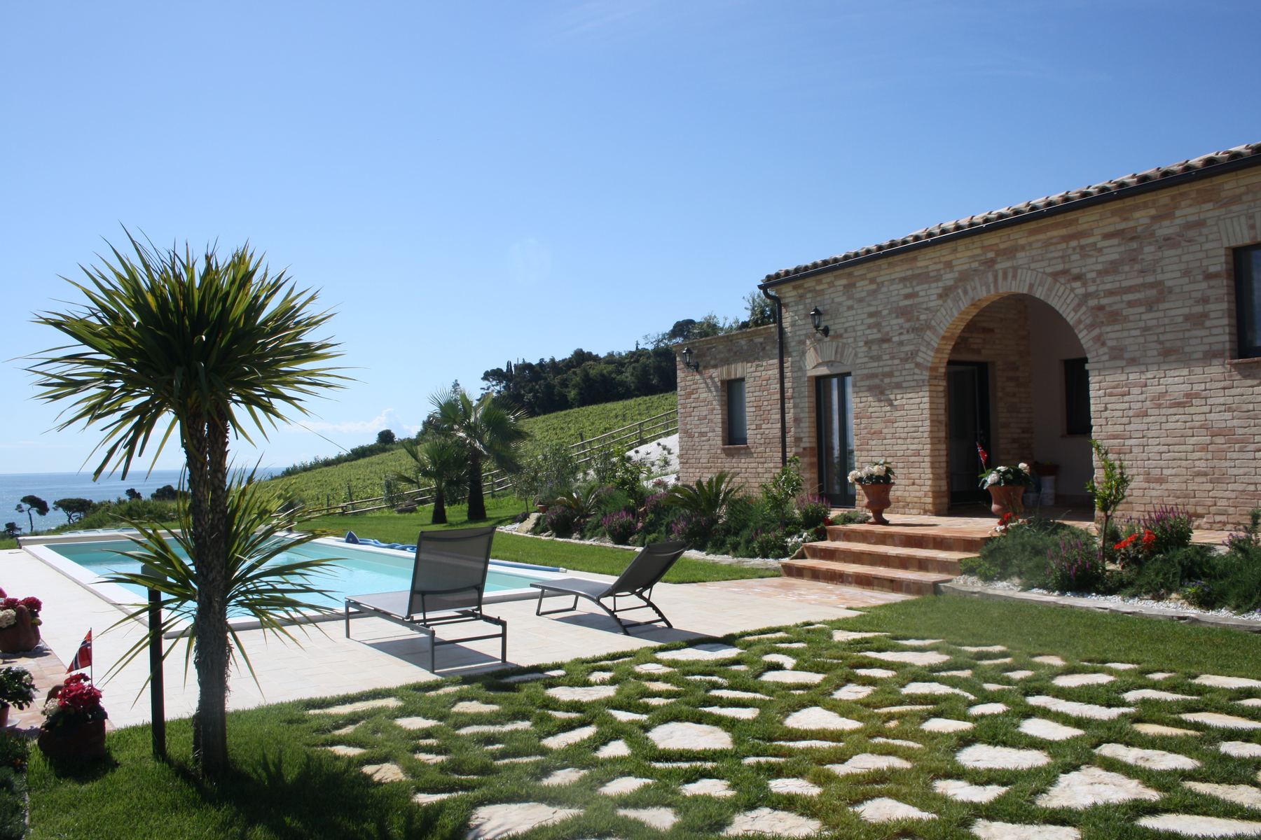 Corte con piscina, lettini e pavimentazione in pietra davanti a un fronte in pietra con grande arco, vista mare